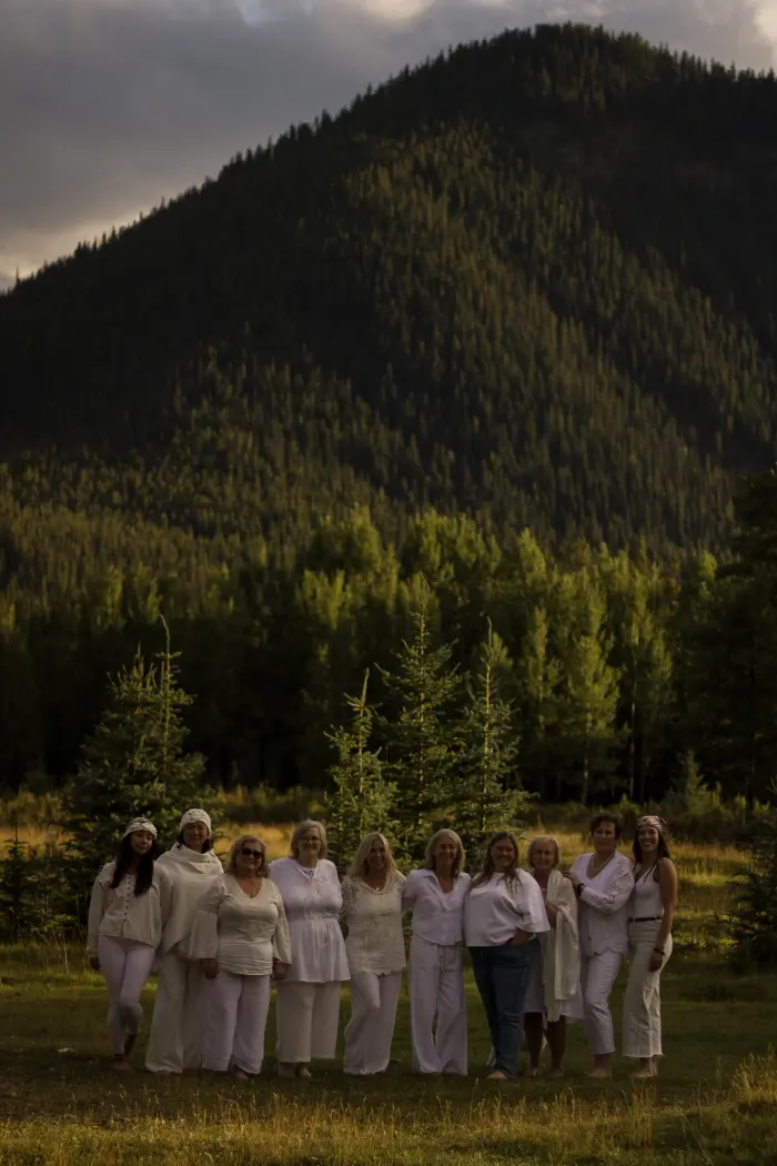 A group of women dressed in white stand together in a meadow below a forested mountain