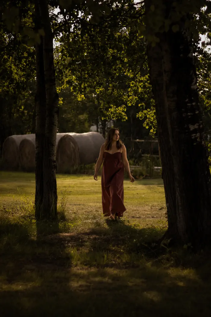 A young woman in a long rust dress walks between two big trees with hay bales behind her
