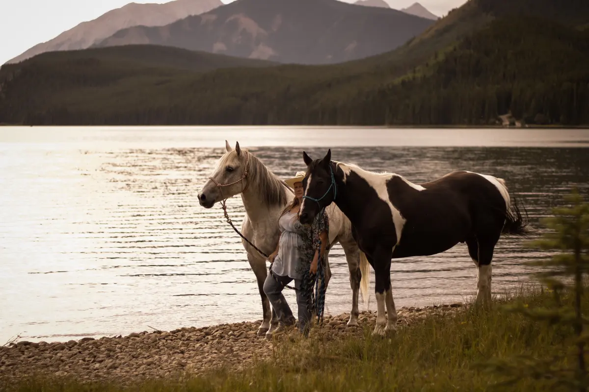 A woman stands between a grey horse and a paint horse on a pebbled lakeshore below the mountains