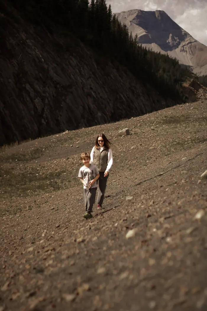 A mother and her young son walk down a rocky slope with a mountain peak behind them
