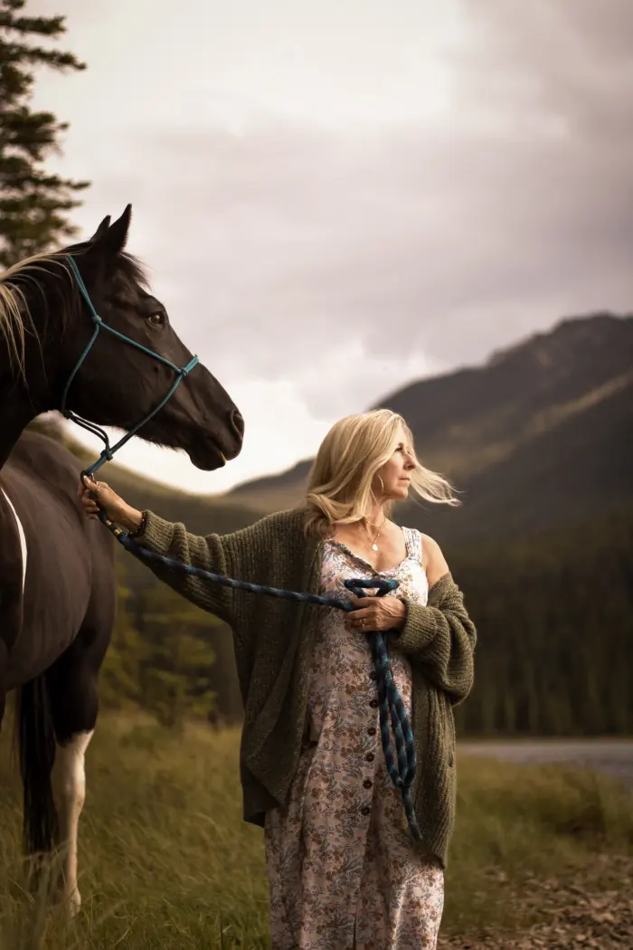 A woman in a floral dress and knit cardigan stands beside her paint horse below the mountains