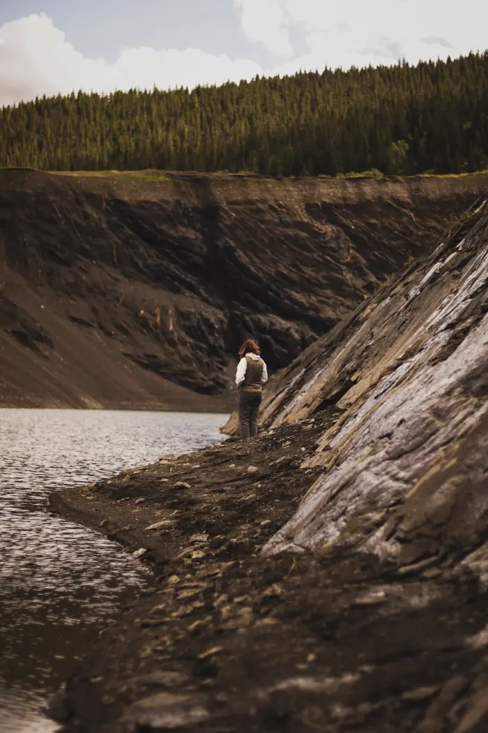 A person stands at the edge of a lake beneath a tall wall of grey shale