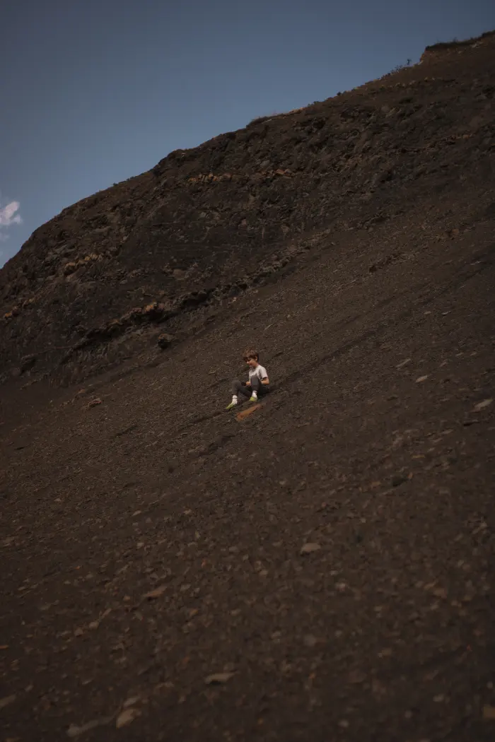 A small boy slides down a huge dark shale slope under a blue sky