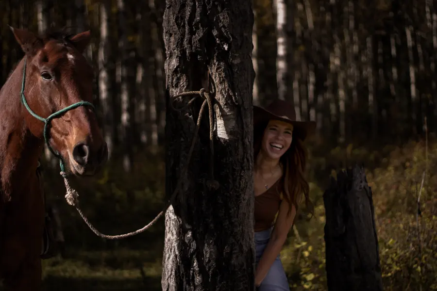 Trudy laughing as she peeks out from behind a big tree, her horse tied close by
